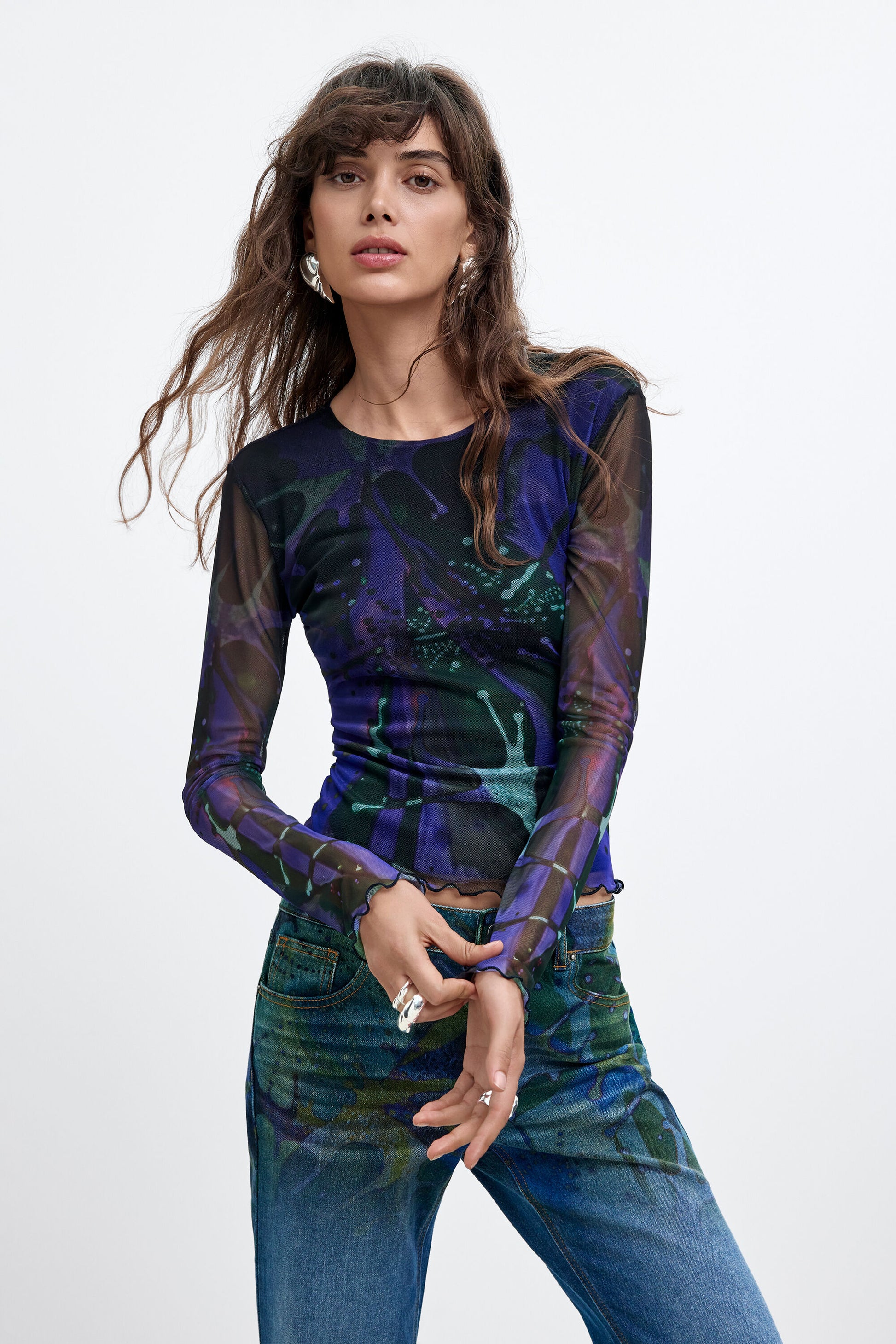 A woman modeling a sheer long-sleeve top with a dark blue, purple, and green abstract print, paired with blue jeans, against a plain light gray background.